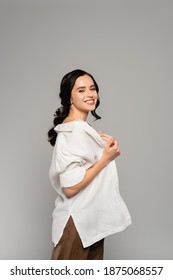 Cheerful Brunette Woman Looking At Camera While Taking Off White Shirt Isolated On Grey