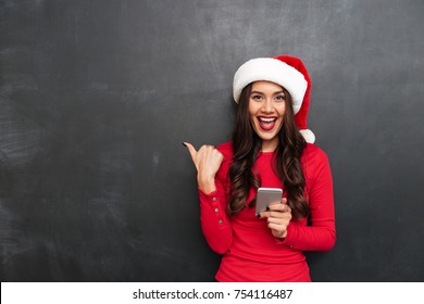 Cheerful Brunette Woman In Red Blouse And Christmas Hat Holding Smartphone And Pointing Away While Looking At The Camera Over Black Background