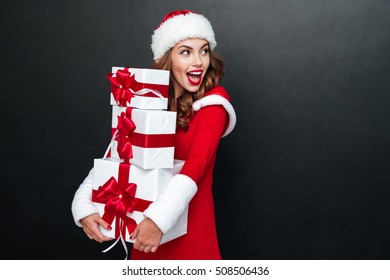 Cheerful brunette woman in red santa claus outfit holding xmas presents isolated on the black background - Powered by Shutterstock