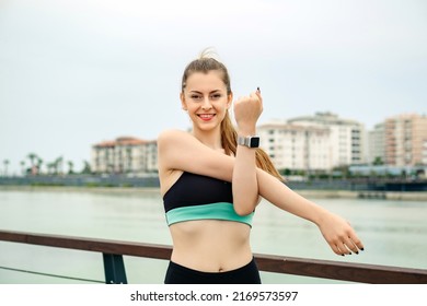 Cheerful brunette woman wearing sportive clothes on city park, outdoors stretching her arms. Female doing warmup stretching workout while looking at the camera.