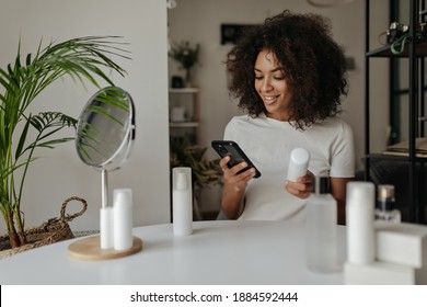 Cheerful brunette woman in white t-shirt smiles, reads recommendations in smartphone and holds great face cream bottle. Lady poses at home.
