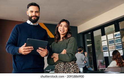 Cheerful Business Colleagues Smiling At The Camera. Two Young Businesspeople Standing In A Boardroom With Their Colleagues In The Background. Diverse Entrepreneurs Working Together As A Team.