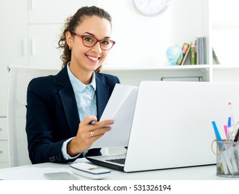 Cheerful business lady sitting at office desk with laptop