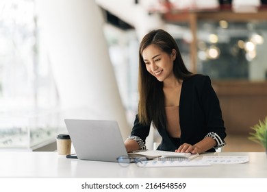 Cheerful business lady working on laptop in office, Asian happy beautiful businesswoman in formal suit work in workplace. Attractive female employee office worker smile.