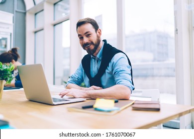 Cheerful business man in blue t shirt smiling and looking at camera while sitting at table and working on laptop in conference room