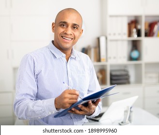 Cheerful business man in light shirt posing in office holding clipboard with papers document write notes