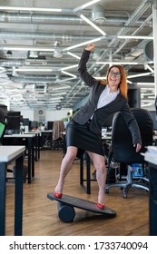 Cheerful Business Woman In Red High Heel Shoes Lost Equilibrium On The Balance Board And Falls. A Female Employee In A Suit Does Sports At The Workplace During The Break. Bad Coordination.