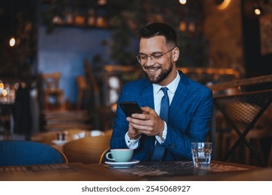 A cheerful businessman in a blue suit and glasses is sitting in a café, engaging with his smartphone and enjoying a coffee. The setting radiates warmth and relaxation. - Powered by Shutterstock