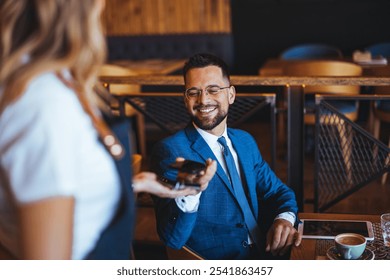 A cheerful businessman in a blue suit uses his smartphone to make a payment in a trendy cafe. He is smiling, engaged and enjoying the modern convenience of technology. - Powered by Shutterstock