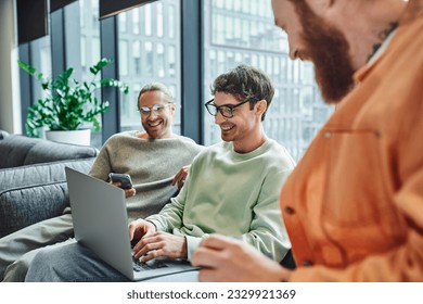 cheerful businessman in eyeglasses typing on laptop while sitting near colleague with mobile phone and bearded man on blurred foreground in lounge of modern coworking space - Shutterstock ID 2329921369