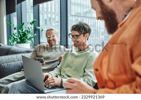 cheerful businessman in eyeglasses typing on laptop while sitting near colleague with mobile phone and bearded man on blurred foreground in lounge of modern coworking space