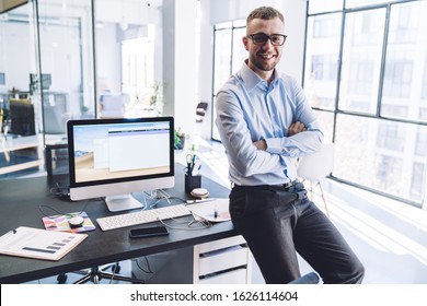 Cheerful businessman in glasses dressed in formal clothes standing with hands crossed near desk with computer in office and smiling at camera - Shutterstock ID 1626114604