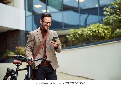 A cheerful businessman with glasses on reading text messages on a smart phone and pushing a bicycle. - Powered by Shutterstock