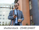 A cheerful businessman stands outside, holding a coffee cup and smartphone, enjoying a break. He wears a suit and glasses, exuding confidence and positivity in an urban setting.