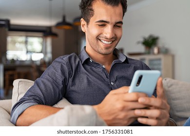 Cheerful Businessman Using Smartphone While Sitting On Sofa At Home. Handsome Young Indian Man Sitting On Couch Reading Messages On Mobile Phone. Hispanic Guy Working From Home With Cellphone.