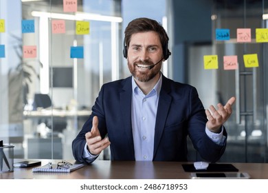 Cheerful businessman wearing headset and talking during a video conference in a modern office. The background features colorful sticky notes on a glass wall. Concept of communication - Powered by Shutterstock