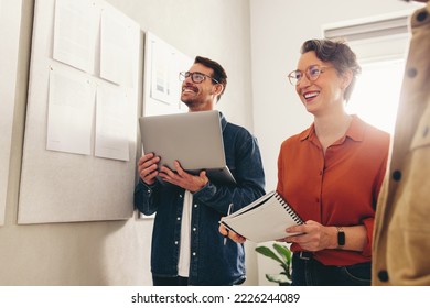 Cheerful Businesspeople Smiling Happily While Having A Meeting In A Creative Workplace. Group Of Business Colleagues Analysing Some Marketing Reports On A Notice Board.