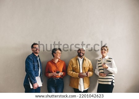 Cheerful businesspeople smiling happily while waiting in line for an interview. Group of shortlisted job applicants holding different digital devices in a modern office.