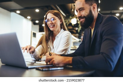 Cheerful businesspeople using a laptop in an office. Happy young entrepreneurs smiling while working together in a modern workspace. Two young businesspeople sitting together at a table.