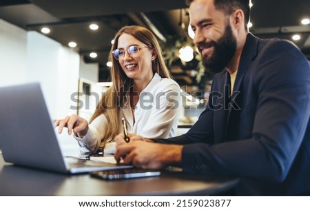 Cheerful businesspeople using a laptop in an office. Happy young entrepreneurs smiling while working together in a modern workspace. Two young businesspeople sitting together at a table.