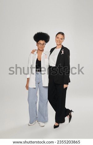 cheerful businesswoman hugging african american woman with pink ribbon on grey, breast cancer month