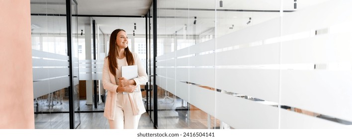 A cheerful businesswoman in an ivory suit walks through a modern glass office corridor, carrying a laptop, her smile suggesting a successful meeting or a job well done, panoramic view, banner - Powered by Shutterstock