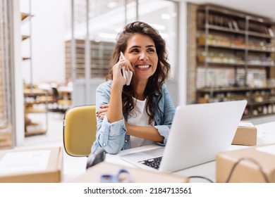 Cheerful businesswoman speaking on the phone while working in a warehouse. Happy online store owner making plans for product shipping. Female entrepreneur running an e-commerce small business.