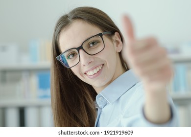 Cheerful businesswoman thumbs up, she is posing in the office and smiling at camera, achievement and satisfaction concept