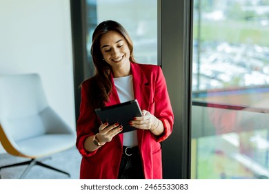 Cheerful businesswoman wearing a red blazer stands by a large window in a modern office, engaging with her tablet as natural light streams in - Powered by Shutterstock