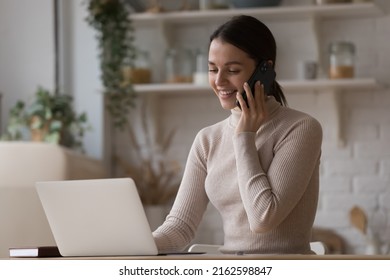 Cheerful busy freelance professional woman working at home kitchen table, speaking on cellphone, typing on laptop computer, laughing, giving consultation. Distant job, communication concept - Powered by Shutterstock