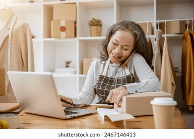 A cheerful, busy mature Asian female tailor or online clothes shop seller is multitasking, talking on the phone with her customer while working on her computer in her atelier studio. - Powered by Shutterstock