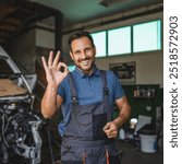 A cheerful car mechanic in a blue polo and overalls gives an okay sign while standing next to a car in a workshop, illustrating positive and professional service