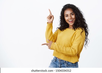 Cheerful carefree african-american woman in yellow sweater with curly haircut, smiling and laughing joyfully dancing as pointing sideways, showing left and up copy space, white background