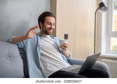 Cheerful carefree guy using laptop computer for video connection, looking at the webcam and waving hand while drinking coffee. Man sitting at the sofa and making video call to somebody - Shutterstock ID 2208336017