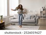 Cheerful carefree young Latin woman dancing in new modern apartment interior, celebrating home buying, property rent, hopping at pale cozy sofa, laughing, enjoying music. Full length