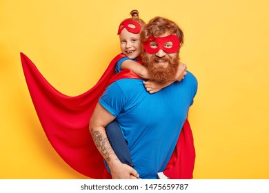 Cheerful Caring Father Plays With Little Daughter Gives Piggyback To Heroic Child, Wear Superhero Suits Have Fun Together While Mommy Is Away Isolated On Yellow Studio Wall. Close Knit Family Concept.