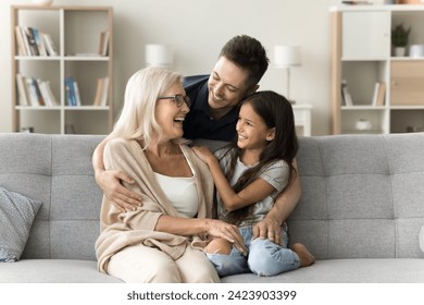 Cheerful caring handsome dad hugging grandma and daughter kid with love, affection on home sofa from behind. Granddaughter, grandmother and adult son enjoying family relationship, talking, laughing - Powered by Shutterstock