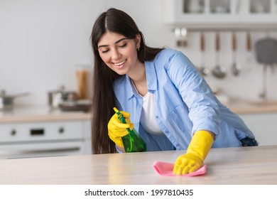 Cheerful casual young woman in yellow rubber gloves cleaning table in cozy white kitchen, using spray and duster cloth. Selective focus, free copy space. House-keeping concept