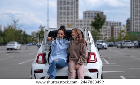 Cheerful Caucasian adult beautiful woman with teen girl kid looking at smartphone camera taking selfie photos sitting in car trunk. Mother and daughter posing to cellphone camera.