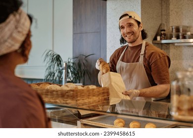 Cheerful caucasian bakery worker putting croissant into craft bag for unknown female client - Powered by Shutterstock