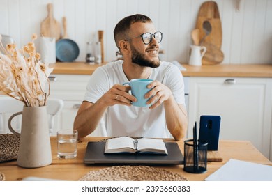 Cheerful caucasian bearded man in glasses sitting at table holds cup of coffee looks away happily, thinking about career, remote works with laptop, planning agenda. Purposeful guy remote working. - Powered by Shutterstock