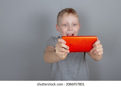 A cheerful caucasian boy looks at his red smartphone and is very surprised on a gray background in the studio.