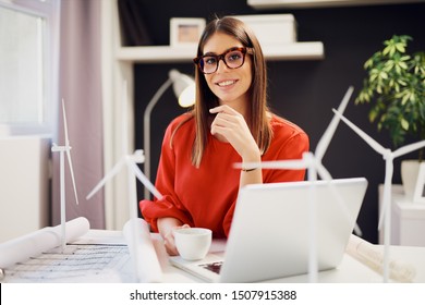 Cheerful caucasian businesswoman dressed in red blouse sitting in office and holding cup with coffee. On table are blueprints, windmill models and laptop. Sustainable development concept.