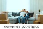 Cheerful Caucasian couple boyfriend and girlfriend moving in together in new house in good mood. Tired happy young married couple sitting down on sofa in new home with many boxes on moving day