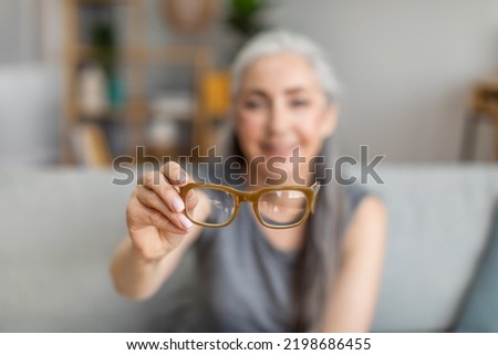 Cheerful caucasian elderly gray-haired female holding glasses in living room interior, selective focus, blurry. Health care, poor eyesight, age-related problems, myopia and advertising, free space