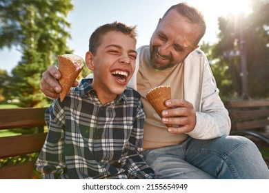 Cheerful caucasian father and teenage son with cerebral palsy eat ice cream and joke on bench in sunny park. Family relationship and enjoy time together. Disability care, treatment and rehabilitation