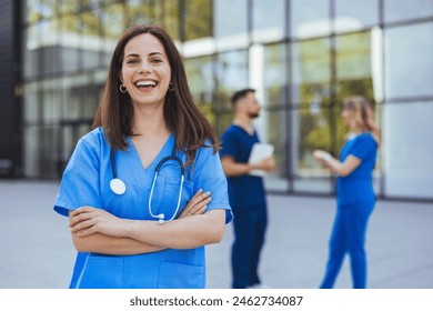 A cheerful Caucasian female nurse, clad in blue medical attire with a stethoscope, stands confidently outside a hospital as colleagues converse in the background. - Powered by Shutterstock