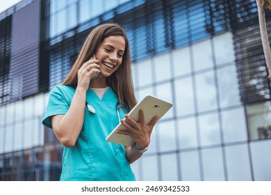 A cheerful Caucasian female nurse in scrubs engages with a smartphone while holding a tablet, echoing the multitasking nature of modern medical professionals outdoors. - Powered by Shutterstock