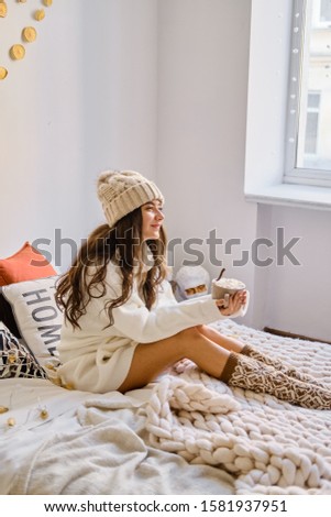  A cheerful Caucasian
 girl in a hat and sweater sits on the bed with a cup of hot chocolate with marshmallows. Cozy interior and New Year's decor.
