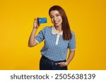 Cheerful caucasian girl holding credit card, recommends bank service, stands over yellow background, laughing and wearing blue shirt